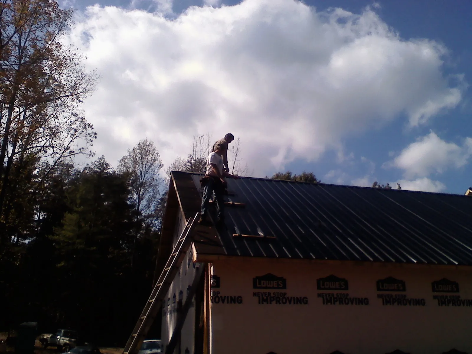 Roofer fastening dark metal panels high against a cloudy sky, from a Roof Inspection job in Flora